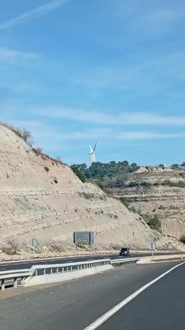 The video shows a highway driving scene. The road is mostly straight with some gentle curves. The landscape features hills with layered rock formations and wind turbines in the background. Cars and trucks are visible on the highway, moving in both directions. The sky is clear with a few wispy clouds.