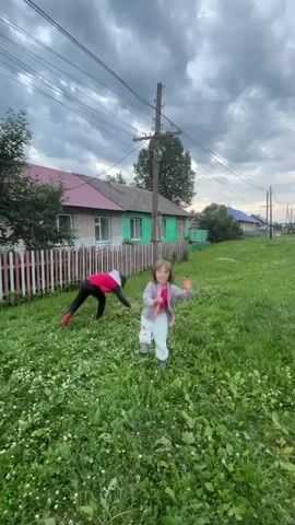 Two children are playing in a grassy area with small white flowers, possibly clover. In the foreground, a younger child with light brown hair, wearing a light-colored top and light blue pants, runs towards the right. Further in the background, a second child, wearing a red top and dark pants, is either falling or getting up from the ground. The children are in a village setting, with a row of houses visible behind a white picket fence. The houses are modest, with light-colored walls and pink or blue roofs. Power lines run across the sky, supported by wooden poles. The sky is overcast with dark, dramatic clouds, suggesting it might be late afternoon or evening, possibly before or after a storm. The overall atmosphere is one of casual, outdoor play in a rural environment. There is no visible text in the media file.