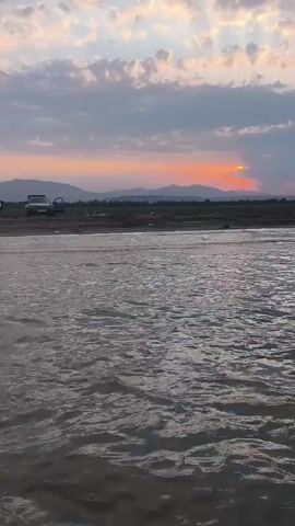 A woman with long, dark red hair walks into a shallow river. She is wearing a dark-colored swimsuit. The water is rippling and brown. In the background, there are hills, a few parked vehicles, and a cloudy sky with the sun setting in the distance. The scene appears to be a natural landscape.