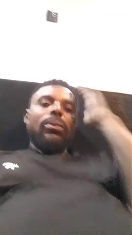 A man with dark skin and a beard is speaking into the camera. He is wearing a black t-shirt with a white logo on the left side of his chest. He is sitting down, with his right arm raised and his hand on his forehead. The background is a white wall with a dark sofa or chair behind him and a small black electronic device mounted on the wall above him.