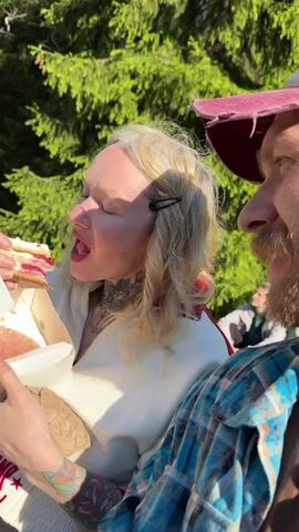 A fair-skinned woman with blonde hair is eating a pastry. The pastry appears to have a red filling. She is holding a white box in her left hand. Her hair is held back by a black barrette. She is wearing a white top with a red pattern around the collar. To her right is a man with a red beard. He is wearing a plaid shirt and a red hat. The background is a green lawn and trees. Other people can be seen in the background.