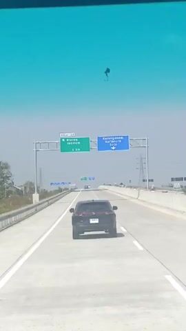 A car is driving down a highway. Overhead signs indicate that the exit for Klaten Jatinom is 1 km away, and Karanganyom Kartasura Solo is in the direction of the arrow. The sky is bright and hazy. There are utility poles visible in the distance.