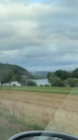 The video shows a landscape with a body of water, trees, and fields. The sky is cloudy. There is a stubble field in the foreground. In the middle ground, there is a body of water with trees along its banks. In the background, there are hills and some distant buildings. The camera is moving, suggesting it is from a vehicle.