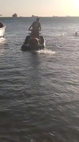 The video shows a man riding a dark-colored jet ski on the water, heading away from the camera. In the background, several large ships are visible on the horizon. The water appears choppy, and the jet ski is creating a wake behind it. There's another smaller boat to the left. The sky is bright, suggesting it could be daytime. The sun is causing glare on the water's surface.