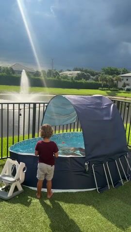 A father and his two young children are enjoying an inflatable pool in their backyard. The father is sitting in the pool with one child, while the other child stands on the grass looking at them. The pool is covered by a blue canopy, providing shade from the sun. In the background, there is a lake with a fountain and houses. The sky is partly cloudy with a ray of sunlight shining through.