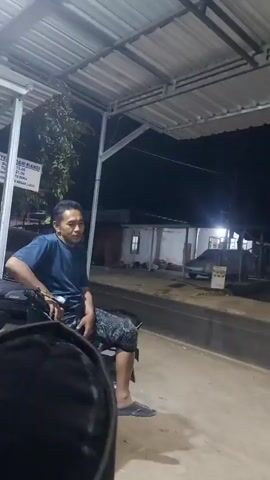 A man in a blue t-shirt sits on a motorcycle under a covered structure. He is gesturing with his hands and appears to be speaking. In the background, there are buildings, a car, and a street. The lighting suggests it is nighttime.