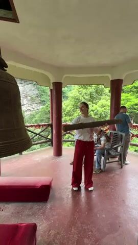 Inside a pagoda-like structure with a red floor and red columns, a woman in a white shirt and red pants stands, holding a large wooden log. She swings the log to strike a large, bronze bell suspended from the ceiling. The bell is engraved with what appear to be Chinese characters. Behind the woman, people are seated on chairs. Through the open sides of the structure, a view of a lush, green mountainside is visible. Red cushions are placed on the floor of the pagoda.
