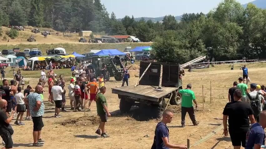 The video shows an outdoor event with a large gathering of people. In the foreground, several men are standing around a dirt area. To the left, a group of people are clustered together, watching something. In the center of the frame, a large, flatbed truck with wooden sides is parked on the dirt. The truck appears to be on an incline and is tilted slightly. A green tractor is positioned behind the truck, and its front loader is raised. There are several tents set up in the background, with more people milling about. Beyond the tents, a wooded hillside is visible, and in the distance, there are mountains. The sky is clear and sunny.
