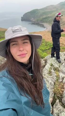 A woman with long brown hair is recording a selfie-style video on a cloudy day. She is wearing a grey sun hat, a black turtleneck, and a teal raincoat. She is smiling and looking at the camera. In the background, there is a man wearing a black jacket and pants, and another person wearing a red hat and a tan jacket. They are standing on a grassy, rocky hill overlooking a body of water and a sandy beach. The woman moves the camera, showing more of the scenery.