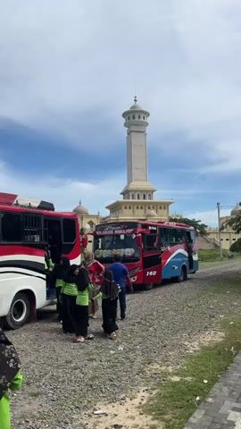 The video shows a group of people near two buses, with a tall minaret and a building in the background. The minaret is white and octagonal at the top, with a dome. The building has a light yellow facade with arched windows and several domes. The buses are red and white, with "JETBUS" written on them. Some people are wearing hijabs and traditional Islamic attire. The ground is covered with gravel and some grass. The sky is blue with scattered clouds.