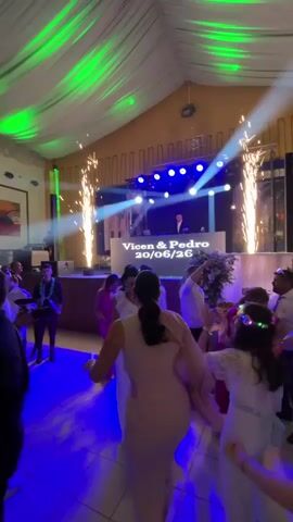 The video shows a wedding reception with a DJ booth in the background. The DJ booth has "Vioen & Pedro 20/06/26" written on it. There are pyrotechnics going off on either side of the DJ booth. Several people are dancing on the dance floor, which is lit with blue and green lights. One woman in a light-colored dress is dancing in the foreground. Another woman with a pink dress is dancing to her left. A man in a white shirt is dancing in the background. The ceiling is decorated with green fabric and lights. There are also some string lights.