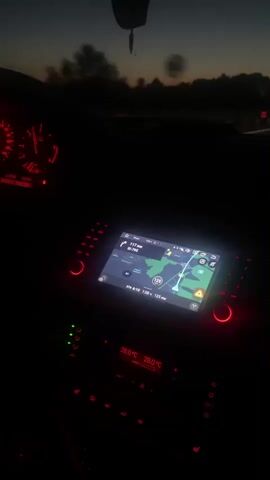 The video begins inside a car at dusk. The dashboard lights are on, and a navigation screen is visible, showing a route. The camera then pans to the windshield, revealing a highway with the sky showing colors of sunset or sunrise. There are orange reflectors on the side of the road. The camera continues to look forward, showing the road ahead as the car travels. The scene transitions between the interior of the car and the view of the highway. There are no people clearly visible.