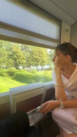 A woman sits in a train car, looking out the window at the passing scenery. She has her chin resting on her hand and her elbow on the armrest. She is wearing a white t-shirt, a watch, and bracelets. The train car is clean and modern, with a large window. The scenery outside is blurred due to the speed of the train, but appears to be a green, tree-filled landscape.