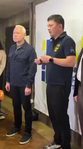Two men stand in front of a backdrop. The man on the left has grey hair and is wearing a dark blue button-down shirt and dark pants. The man on the right is wearing a dark blue polo shirt with a logo on the chest, dark pants, and a watch. He is gesturing with his hands as he speaks. A third person is partially visible to the right. The backdrop is white with blue and yellow rectangles. The room appears to be indoors.
