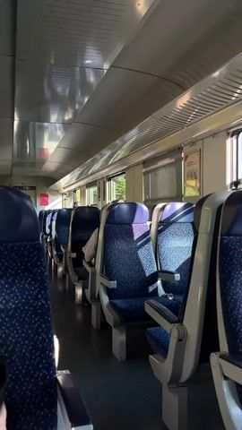 The video shows the interior of a train carriage. The seats are blue with a speckled pattern and have silver armrests and headrests. They are arranged in rows on either side of an aisle. Sunlight streams in through the windows, casting shadows on the seats and the floor. One person is visible, seated and facing away from the camera, partially obscured by the seat in front. The ceiling of the carriage is metallic and reflects the light. There are overhead compartments above the seats and windows along the side. Advertisements or information posters are affixed to the wall between the windows. The camera appears to be stationary, looking down the length of the carriage.