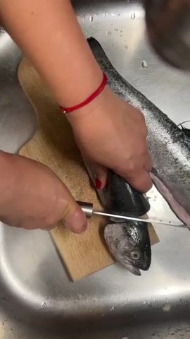 A top-down view shows a person with a red bracelet and red nail polish, holding a silver knife, cutting the head off a fish in a stainless steel sink. The fish has silver scales and is lying on a small wooden cutting board. Another similar fish lies to the right of the first fish. The person's left hand holds the fish body, while their right hand uses the knife to sever the head, which is then separated from the body. The cutting board is rectangular with rounded edges.
