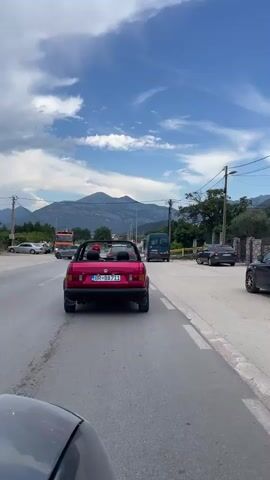 This media file captures a scenic drive on a sunny day, likely in Tomba, Montenegro. The primary focus is a vibrant red convertible Volkswagen Golf Cabriolet, seen from behind as it travels along a paved road. The car's license plate reads "BR BA 711". Inside the convertible, two figures are visible, one with a red helmet, suggesting they are enjoying a leisurely drive.

The setting is a semi-urban or rural landscape, with mountains forming a majestic backdrop. The sky is a brilliant blue, dotted with fluffy white clouds, indicating pleasant weather. Along the roadside, other vehicles are present, including an orange truck on the left and a green van further ahead, as well as parked cars and what appears to be a small roadside establishment. Power lines and streetlights are also visible, typical of a road in a populated area. The road itself is asphalt, with white lane markings. The overall atmosphere is relaxed and suggests a pleasant journey through a picturesque location.