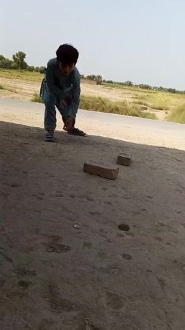 A young boy wearing a light blue shalwar kameez is crouching on a dirt ground, reaching for something near two bricks. He is in front of a shaded area, with a blurred background of a dry, grassy landscape under a bright sky. Another person, partially visible from the waist down wearing light-colored pants and gray sandals, is entering the frame from the right. This second person is also crouching and appears to be interacting with the boy or the objects on the ground. The focus is on the ground and the two individuals, with the environment serving as a backdrop.