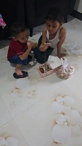 Two young boys are sitting on a patterned tile floor, eating snacks. The boy on the left is wearing a red shirt and blue shorts, and is wearing black sandals. He is holding a snack in his hand and eating it. The boy on the right is wearing a white tank top and green pants. He is holding a snack in his hand and eating it. In front of the boys is a small pink and white patterned box containing more snacks, and a clear plastic bag full of round snacks. The floor has a floral pattern.