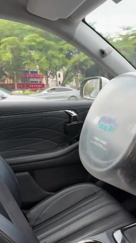 The video shows the interior and exterior view of a vehicle driving on a city street during daylight.  The interior of the car is black with quilted leather on the door and seats. A large, round, metallic-looking object is mounted on the passenger seat.  The car's dashboard has a large digital display showing a navigation app indicating the vehicle's current location and route. The screen shows "Remaining 4.5 km Arrival in 10 min."  A dashcam is visible above the screen. The car is driving on a multi-lane road with other vehicles. Outside, buildings and trees line the street.