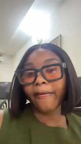 A medium-shot video shows a woman with brown skin and a black bob-cut hairstyle. She wears black framed glasses with orange-tinted lenses. She is also wearing an olive-green top. She subtly moves her head. Behind her is a television, a framed picture, and a security camera. The room has a white ceiling with recessed lighting.