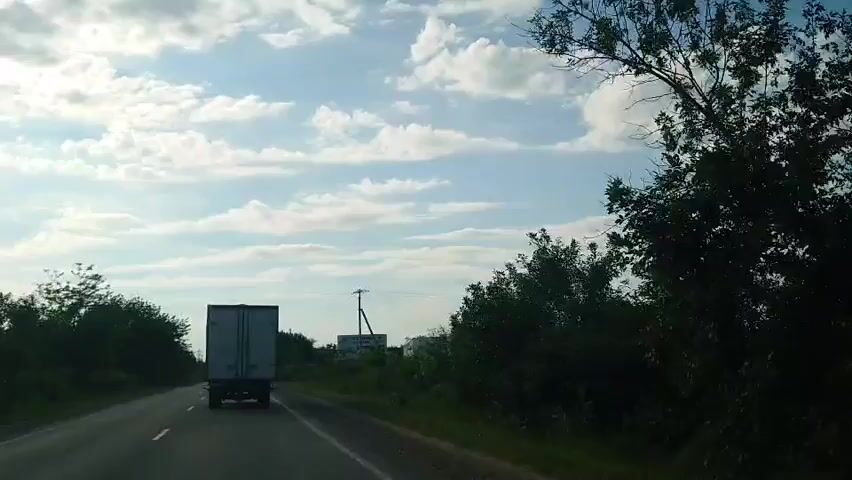 A rear view of a white truck with a grey trailer driving on a paved road. Trees line the left and right sides of the road. A utility pole with wires is visible on the right side of the road, next to a billboard. The sky is blue with scattered white clouds. The video is shaky and appears to be taken from a moving vehicle.