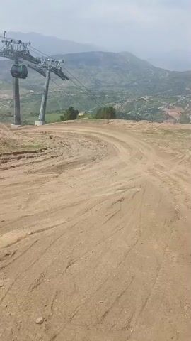 The video shows a dirt road with tire tracks leading away from the camera. In the background, there is a cable car and a mountainous landscape. The sky is overcast. The camera appears to be mounted on a vehicle, as indicated by a wiper blade visible at the bottom right of the frame. The overall scene is of a rural, possibly mountainous area.