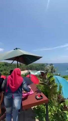 The video shows a scenic view of a coastal camping site on a sunny day. Several tents, red, blue, and gray, are pitched on a grassy area overlooking the ocean. A concrete path runs through the camping site, and people, including a man with a child, are seen walking along it. In the foreground, there is a wooden table with a tray holding drinks and snacks. A woman in a red hijab stands by the table. An umbrella provides shade. The ocean has rocky formations and waves crashing against the shore. The sky is clear and blue with a few scattered clouds. Green trees surround the area, adding to the natural beauty of the location.