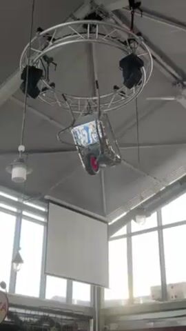 The video shows a reflective, disco ball-covered wheelbarrow hanging from the ceiling of a building, suspended by chains. The wheelbarrow has a red wheel. Above the wheelbarrow is a circular lighting rig with several spotlights attached. A projection screen hangs nearby, and there are some windows in the background. The ceiling is a light grey. There is music playing in the background.
