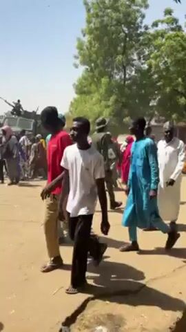 The video shows a group of people walking on a dirt road on a sunny day. The individuals are diverse in age and wear a variety of clothing.  Several are dressed in traditional attire, including long white robes. A child in patterned clothing carries a flat, round object. The group appears to be moving with purpose. Trees and buildings are visible in the background.
