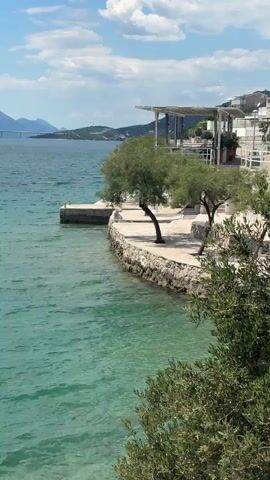 The video shows a beautiful view of a turquoise sea with a rocky coastline. The sky is blue with scattered white clouds, and the sun is shining. Along the coastline, there are trees and buildings, including a white pergola structure. In the distance, a bridge spans across the water to a hilly landmass. The camera pans slightly, revealing a stone pathway and more vegetation, suggesting a coastal area.