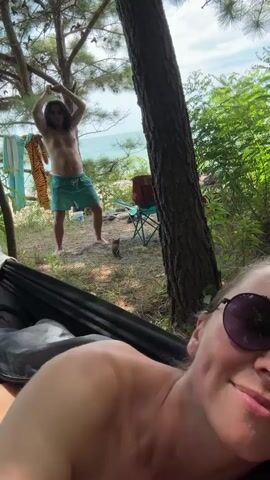 The video begins with a close-up, low-angle shot of a woman in a hammock. She is wearing sunglasses and smiling at the camera. Her shoulder and chest are visible. In the background, a man with long dark hair and a bare chest is standing on a sandy area near trees and the ocean. He is wearing blue patterned swim shorts and appears to be dancing or posing, with his arms raised above his head. Two folding chairs and some towels are visible behind him. The environment is a wooded area on a beach. The video is a short selfie-style clip, with the camera focused on the woman in the foreground and the man in the background slightly out of focus.