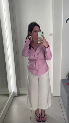 A young woman with brown hair pulled into a ponytail and wearing a pink off-the-shoulder button-down shirt with a twisted knot in the front and white wide-leg pants takes a selfie in a brightly lit dressing room. She is holding a light green smartphone in her right hand. Her left hand is tucked into her pants pocket. She is wearing black flip-flops. The dressing room has a tiled floor and mirrors along one wall. A gray bench with a dark bag on it is visible in the background.