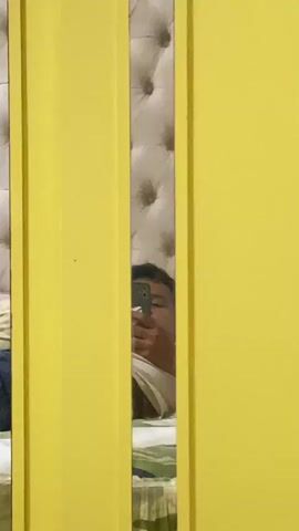 A person is reflected in a mirror. The person is holding a phone and appears to be taking a selfie. The person's face is partially visible. The background of the reflection shows a tufted headboard and a bed with striped sheets. The foreground of the video is dominated by two vertical yellow panels.