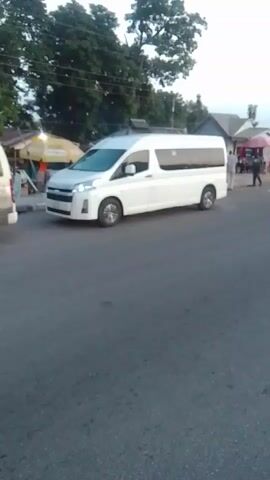 The video begins with a white minibus driving down a paved road. On the left side of the road, there are some market stalls with people standing around them. The minibus passes by these stalls and continues down the road. As the video progresses, other vehicles, including a motorcycle and a silver sedan, are seen driving on the road. The scene appears to be in a populated area, possibly a town or city, with trees and buildings visible in the background. The lighting suggests it is either early morning or late afternoon.