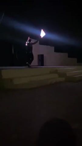 A performer stands on an outdoor stage at night, illuminated by a spotlight from the left. The performer wears a sparkly tank top and dark pants and holds two glowing batons, performing a baton twirling routine. The stage has several tiers of concrete steps leading up to it. There is a speaker on the right side of the stage, and a hoop sits behind the performer. The ground in front of the stage is concrete, and it is cracked. The background is dark, obscuring any specific details. Music plays throughout the video.