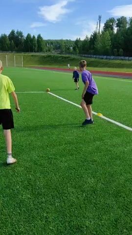 A group of young boys are playing soccer on an artificial turf field. The field is green with white lines marking the boundaries. In the background, there are trees, a fence, and some buildings. One boy is in the foreground, wearing a purple shirt and black shorts, and appears to be kicking the ball. Another boy, wearing a blue shirt and black shorts, is running towards the ball. A third boy, wearing a yellow shirt, is further in the background. The ball is yellow.