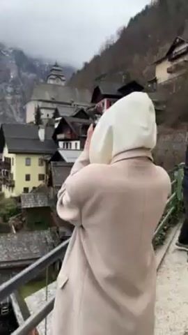 A person wearing a beige coat and a white hood is standing on a bridge, holding a black camera. They are taking a photo of a town nestled in the mountains. The town has a mix of colorful buildings with dark roofs, and a church steeple is visible in the background. The mountain is covered in trees and the sky is overcast.