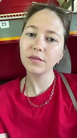 A woman in a red t-shirt and gold chain necklace is looking at the camera. She has blue eyes, fair skin, and her hair is pulled back. Sunglasses are perched on her head. She is sitting in a seat on a train, with red and gray interior panels visible in the background. The number 23 is displayed on one of the panels. She smiles briefly.