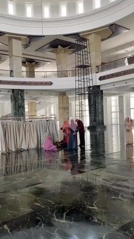 The video shows the interior of a mosque. A large dome with intricate green and yellow patterns is visible at the top of the frame. Below the dome, a tall metal scaffolding structure is erected in the center of the open space. Several people, mostly women wearing headscarves, are gathered on the highly polished, reflective marble floor. One person is kneeling, while others are standing and talking. Another person, dressed in darker clothing, stands near the scaffolding. In the background, there are balconies and large pillars, and natural light streams in through windows. The floor is green and black marble with a reflective surface, showing a mirror image of the people and the structure.