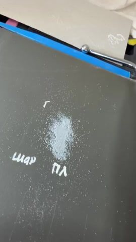 The video shows a close-up of a dark gray surface with a white powdery substance scattered in the center. To the left of the powder, the word "шар" is written in white marker. Below the powder, the letters "ПЛ" are written in white marker. Above the powder, a single letter "Г" is written in white marker. A blue object with a silver metallic part is visible in the upper part of the frame, resting on a beige surface. The beige surface has some faint white markings on it. The audio consists of someone saying "Ну, всё отлично расплавилось," which translates to "Well, it all melted perfectly." The video seems to be documenting a process of melting a substance.