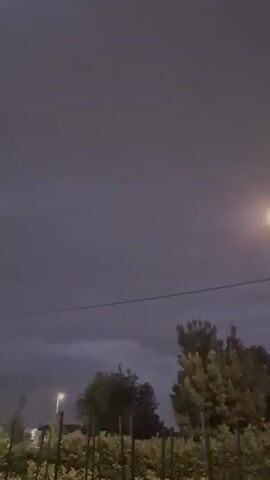 The video shows a slightly cloudy sky at dusk or night. A single street lamp illuminates the scene from the right. In the background, trees and bushes are visible, along with some buildings on the left. A power line stretches across the middle of the frame. The overall atmosphere is calm, with no people or action visible.