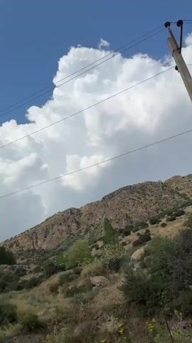 The video shows a landscape with a mountain range in the background under a blue sky with fluffy white clouds. There are power lines strung across the sky. The foreground is a rocky, dry hillside with sparse vegetation. There are no people or visible animals in the video. The camera appears to be moving, possibly from a vehicle, as the perspective shifts slightly throughout the short clip. The terrain suggests a rural or mountainous environment.