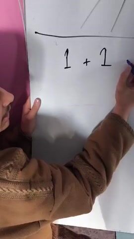 A person in a brown sweatshirt stands in front of a whiteboard, on which is written the equation "1 + 1 = ?" The person is holding a blue marker, then they put their hand on the board. After that, they continue writing on the board. A second hand can be seen touching the board. A pink wall is visible in the background.