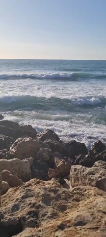 The video captures a serene coastal scene on a bright, sunny day. The camera is positioned from a slightly elevated vantage point, looking out towards the ocean over a foreground of large, rough rocks that form a natural or man-made breakwater. These rocks are varied in size and color, ranging from light sandy brown to darker grays, and appear wet in some areas where the waves have recently crashed.

Beyond the rocks, the vast expanse of the ocean dominates the frame. The water is a beautiful blue-green color, particularly in the deeper areas further from the shore. Closer to the shore, waves are actively breaking and rolling in. Multiple lines of white, foamy waves can be seen. The primary action in the video is these waves crashing against the rocks, sending splashes of white spray into the air, and then receding, leaving behind a foamy residue on the rock surfaces and in the water.

The horizon line is clear and distinct, separating the ocean from a pale blue sky, which appears cloudless. The sun, though not directly visible, casts a bright light across the scene, highlighting the tops of the waves and creating a glistening effect on the water. The overall impression is one of natural beauty and the powerful, continuous movement of the ocean.
