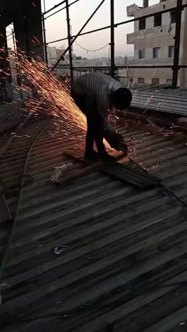 A person is using a power tool to cut a piece of metal on a corrugated metal surface. Sparks are flying from the point of contact. The person is bent over the work, wearing dark pants and a light-colored shirt. The scene appears to be on a rooftop or high platform under a metal frame structure, with buildings visible in the background against a dusk sky.