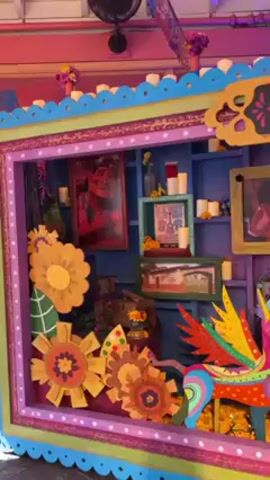 The video shows a brightly decorated display with the words "Recuérdame" in the center. The display features pictures, flowers, candles, and a colorful winged alebrije. It has a purple, blue, and green color scheme. The background music is Mexican folk music.