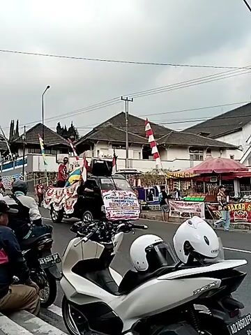 The video shows a parade-like procession on a street in Tawangmangu, Indonesia. The main focus is a black pickup truck decorated with red and white flags and a banner that reads "HADIRILAH DAN SAKSIKAN TURNAMEN VOLLY BUPATI CUP 2025". The truck is carrying several people, some wearing red shirts, and a large colorful inflatable object. There are also several motorcycles in the foreground, one with two white helmets on the seat, suggesting it is part of the procession or spectators. In the background, there are buildings with tiled roofs and some vendors with umbrellas. The weather appears to be overcast, and the time of day seems to be daytime. The atmosphere appears festive, with people participating in or observing the event. The presence of the Bupati Cup banner indicates a sporting event is being promoted.