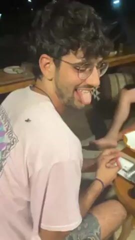 A person with curly hair, glasses, and a pink t-shirt is sitting at a table. A large insect is on their back. The person sticks out their tongue and laughs. There are other people at the table. The environment appears to be outdoors at night.