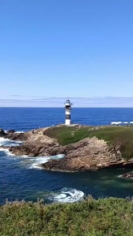 A camera pans across a coastal landscape under a clear blue sky. In the distance, a striped lighthouse stands on a rocky outcrop. The sea is a deep blue, with waves crashing against the rocks. Closer to the camera, there is lush green vegetation. The camera continues to move, revealing more of the coastline and the lighthouse.